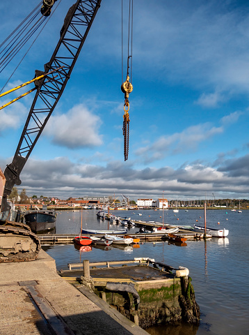 An old crane beside the River Deben at Woodbridge in Suffolk, Eastern England. In the background, the famous Tide Mill can be seen with various leisure craft and house boats, while in the foreground, rowing boats are moored at a jetty.