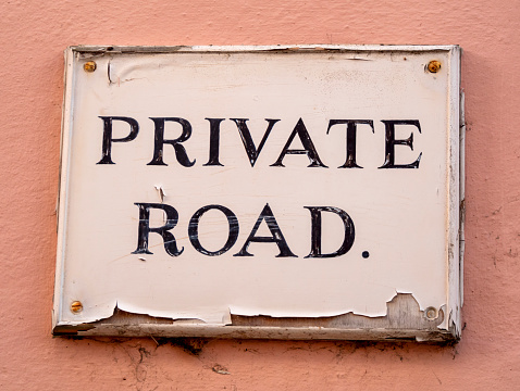 A white-painted and peeling old sign stating ‘Private Road.’ fixed to a coral-coloured wall in Suffolk, Eastern England. The coral pink is generally known as ‘Suffolk Pink’ and is seen all over the county.