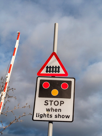 A raised barrier with a warning sign on an unmanned railway crossing in Woodbridge, in Suffolk, Eastern England, on a bright day. Dark clouds are looming, however, with the possibility of rain.