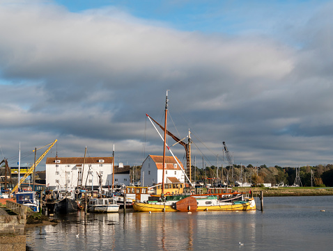 A group of houseboats, sailing barges and other leisure craft moored in Woodbridge, Suffolk, Eastern England, at high tide in the River Deben. The famous Tide Mill can be seen  behind the boats, white against the dark sky which threatens rain.