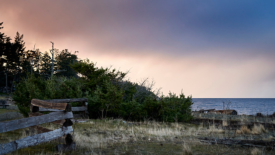 A wooden fence leading the viewer to bushes and the ocean and a colorful sky at dusk