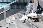 Stylish man in grey jeans and white sneakers put his feet on the longboard. Hipster resting sitting on the steps under the sun.