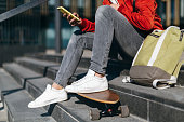 Close up portrait of trendy hipster in red jacket, white sneakers and green backpack with longboard sitting on the steps. Man chatting by the mobile phone.
