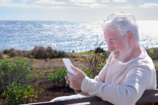 Video call concept. Senior smiling bearded man sitting on a bench face the sea talking on mobile phone using phone webcam to communicate in video chat. Horizon over water