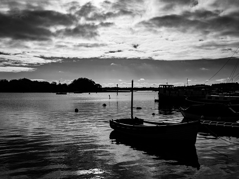 A cluster of boats in deep shadow with a dramatic sky over the River Deben in Woodbridge, Suffolk, Eastern England, late on a sunny November afternoon.