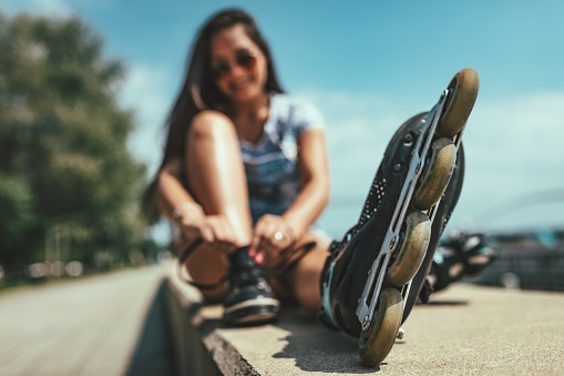 Young female skater is enjoying on the city river shore and ties her rollers on a beautiful summer day. Selective focus.