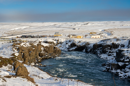 Wild river with cliffs on the banks, covered by snow. Buildings, houses and road in the background. Blue sky with cloud on the horizon. Fossholl - Godafoss, Iceland.