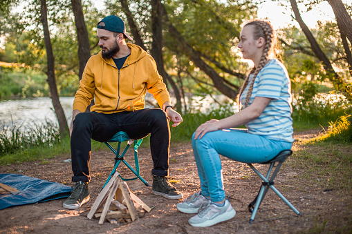 Young hiker man and woman sitting on camping chair and kindling fire in nature