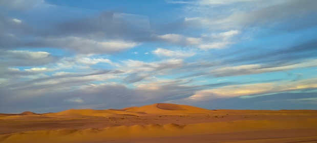 A sweeping panorama captures the vast expanse of golden sand dunes stretching as far as the eye can see, forming an arid desert under a partly cloudy blue sky in Timimoun, Algeria.
