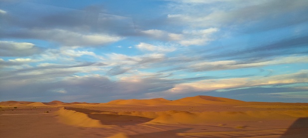 A sweeping panorama captures the vast expanse of golden sand dunes stretching as far as the eye can see, forming an arid desert under a partly cloudy blue sky in Timimoun, Algeria.