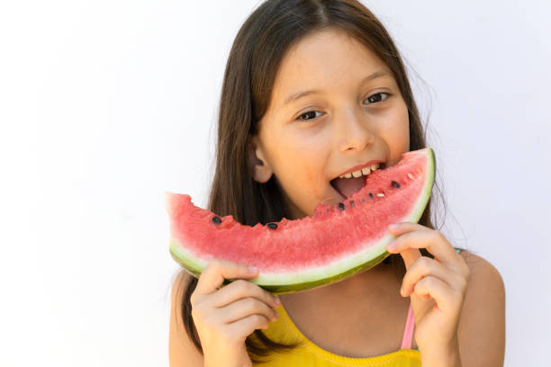 Girl eating a slice of watermelon in front of white background with a cute smile.
