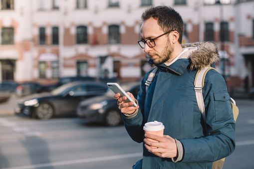 Pleasant male with beard, glasses and in blue jacket looking into mobile phone and drinking coffee. Social media addiction, dependency on a cell phone.