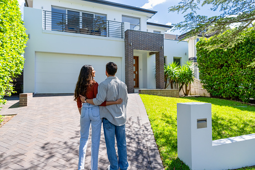 Couple standing in front of their new home. They are both wearing casual clothes and embracing. Rear view. The house is contemporary with a brick facade, driveway, balcony and a green lawn. The front door and mailbox is also visible. Copy space