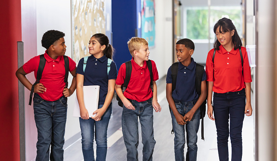 A multiracial group of five middle school students walking together in a school hallway carrying backpacks. They are 11 and 12 years old.
