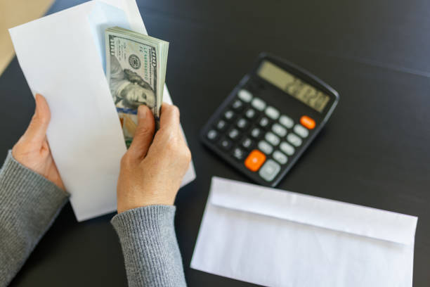 A woman putting a hundred dollar bills into a white envelope. There is a calculator next to her on the table.