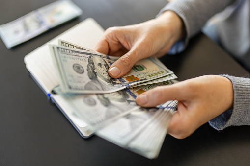 A woman holding a stack of hundred dollar bills. There is a notebook on the table in front of her.