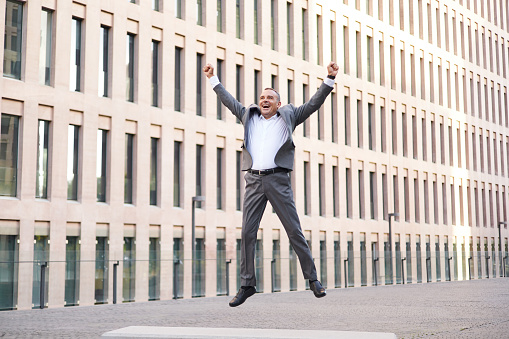 Middle aged male boss in gray suit raising clenched fists and screaming while jumping and celebrating victory outside modern building on city street