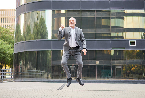 From below middle aged male boss in gray suit raising clenched fists and screaming while jumping and celebrating victory outside modern building on city street