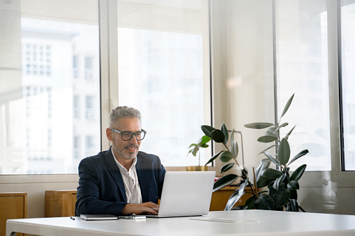 Smiling optimistic mature gray-haired businessman ceo wearing eyeglasses using laptop, responding emails, conducting negotiations online, solving business tasks, planning business strategy