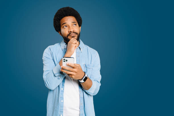 Pensive man appears intrigued as he holds his smartphone, chin resting on his hand Pensive man appears intrigued as he holds his smartphone, chin resting on his hand, conveying a moment of curiosity or deep thought. The concept of problem-solving or information searching. deep pick stock pictures, royalty-free photos & images