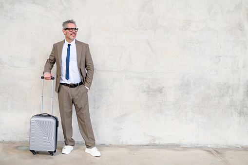 A mature businessman in a smart suit stands with a suitcase, sporting a cheerful smile against a textured concrete backdrop, encapsulating the ease of modern business travel.