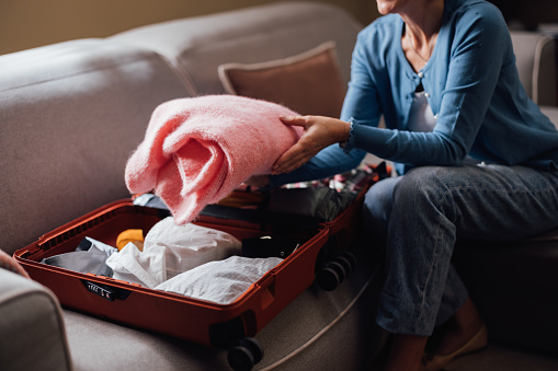 A middle-aged woman in casual clothing neatly packs clothing into an open suitcase, suggesting travel preparation.