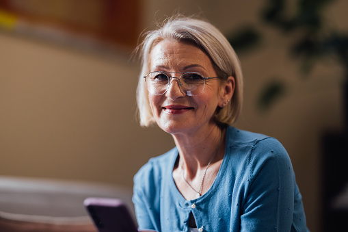 A cheerful senior woman with glasses and a blue top smiles while using her smartphone in an indoor setting.