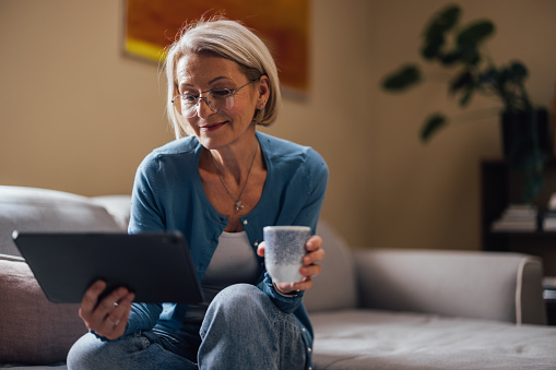An older woman enjoys a peaceful moment on her couch, drinking coffee and browsing a tablet in a cozy living room.
