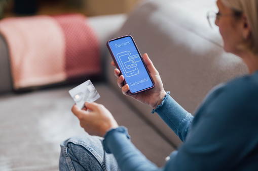 Mature woman holding credit card and smartphone, completing a successful online payment transaction, seated comfortably at home.
