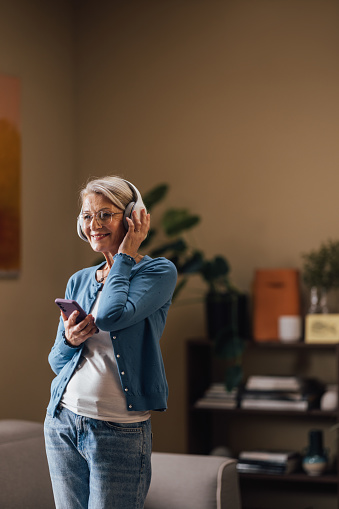 Mature lady with glasses using smartphone and headphones, smiling while standing in a cozy interior.