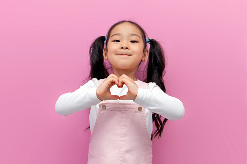 little Asian girl in pink sundress with long hair shows heart with her hands, Korean preschool child makes gesture of love on pink isolated background