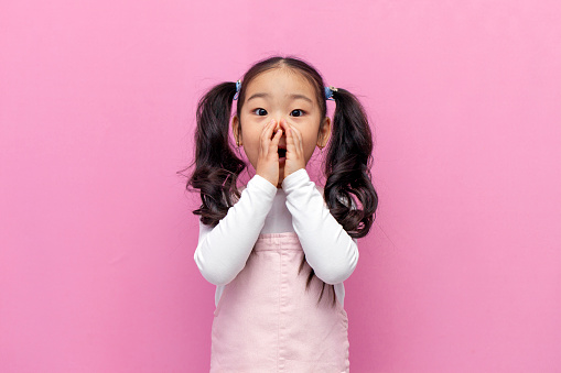 little shocked Asian girl in pink sundress with long hair is surprised and covers her mouth with her hands on pink isolated background, amazed Korean preschool child screams in shock