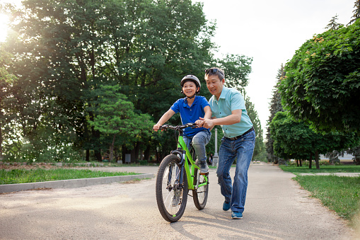 asian dad teaches son to ride bike, korean senior helps child, boy in helmet is actively relaxing with his father in the park in summer