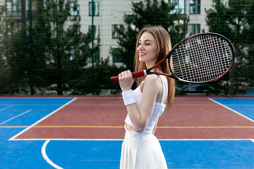 portrait of tennis player girl in white uniform with racket on blue court, female athlete stands near the tennis net and smiles, copy space
