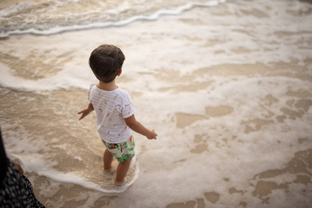Cute little boy standing on the beach in the shallow sea in sunset.