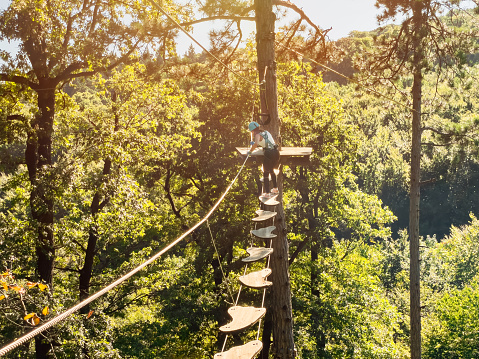 High-altitude climbing training of child on adventure track, equipped with safety straps and protective helmet.Teenage girl goes on hinged trail in extreme rope Park in summer forest.Summer fun and sports for adventurous people.