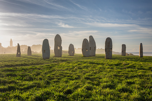As the first light of dawn pierces the clear sky, it reveals an ancient arrangement of menhirs standing solemnly in a lush green field in Galicia. These prehistoric monuments, shrouded in morning mist, whisper tales of Spain's megalithic past, forming a bridge to a time when such stones were erected for reasons that remain a cultural heritage mystery. The grass glistens with dew, and the fog lends an ethereal quality to the landscape, inviting contemplation of nature and history intertwined.