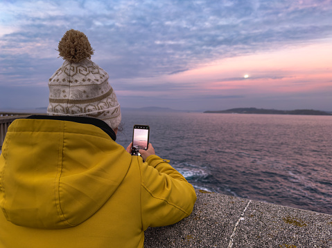 a person clad in a vibrant yellow jacket, their back to the camera, as they capture the moonrise on their smartphone. The subtle moon glows softly in the pastel-hued twilight sky, casting a gentle light over the calm sea. The tranquil scene is a blend of technology and nature, illustrating the modern way of appreciating timeless landscapes.