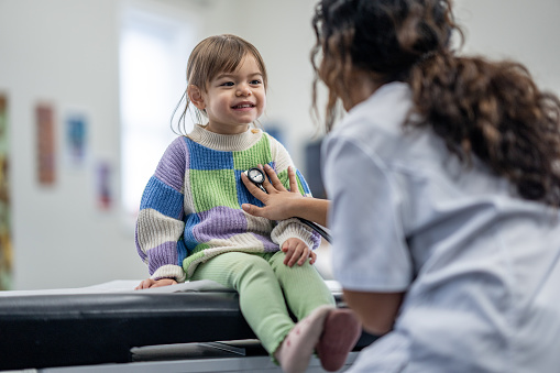 A sweet little mixed race girl sits up on an exam table during a medical appointment.  She is dressed comfortably in a brightly coloured sweater and has a neutral expression on her face as her doctor listens to her heart.