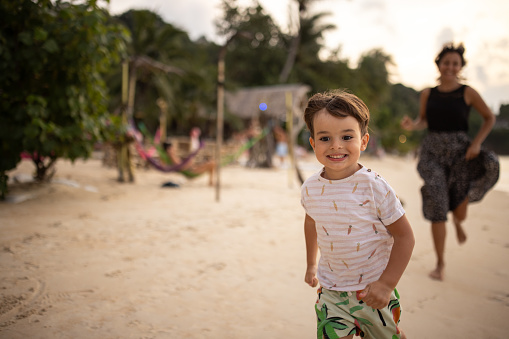 Happy little boy with his mom having fun running on the beach in sunset, they are on vacation in Thailand.