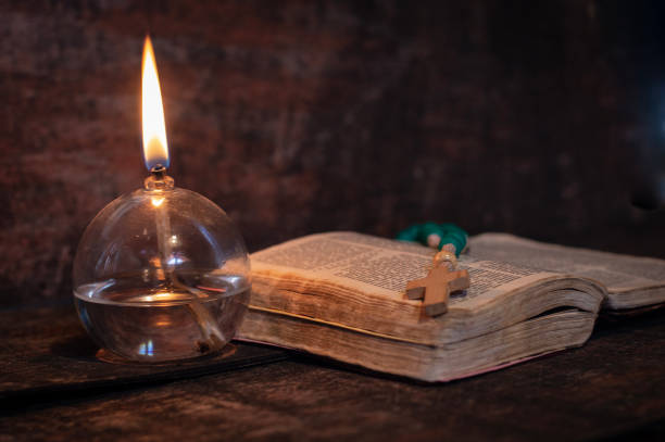 Color rosary on the open bible with a white wooden cross and a lit oil lamp, objects for prayer