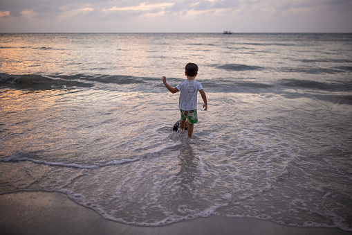 Cute little boy playing on the beach in the shallow sea in sunset.