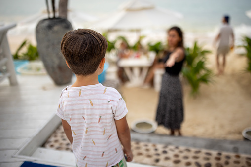Little boy standing at the beach bar, his mother is in front of him, they are on vacation in Thailand.