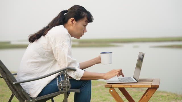 Asian woman typing working on laptop while sitting chair at campsite outdoor.