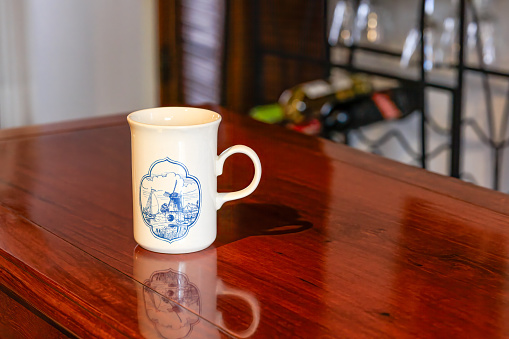 A Coffee Mug Souvenir From Holland placed on a shiny rosewood table. It is an immitation of the typical Delft pottery from the Country, in the typical blue on white porcelain. Image shot indoors in a studio environment. No pepople.