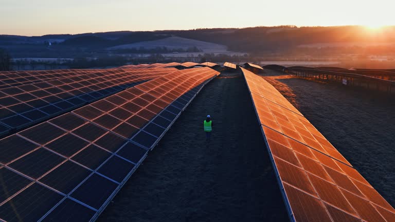 AERIAL Drone Shot of Female Engineer in Reflective Jacket Walking at Solar Power Plant During Sunset