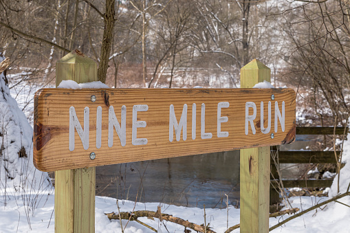 A wooden sign engraved with Nine Mile Run, a stream in Frick Park, an urban park in Pittsburgh, Pennsylvania, USA as seen on a sunny winter day
