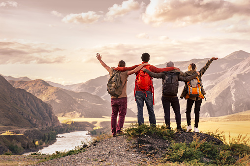 Four happy young tourists with backpacks are standing and embracing in mountains and enjoying sunset