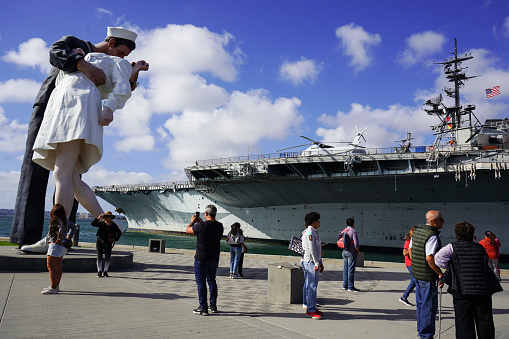 Embarcadero -San Diego, CA ,USA- October 26, 2023.  A lady poses as her photo is taken in front of the Unconditional Surrender Statue.  The Statue is located on the waterfront of the San Diego Bay and is often a photo op. The USS  Midway Museum is in the background.