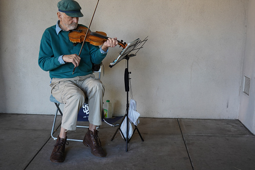 Balboa Park- San Diego, CA ,USA- October 29, 2023. A gentleman street performer sits in a chair, playing his violin in the shade of a hall in Balboa Park.  He concentrates on the stand that holds his sheet music while he taps his feet in time.